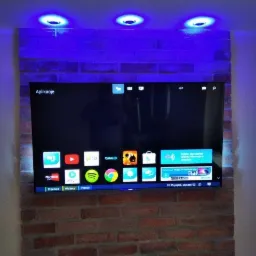 Telewizor LCD zawieszony na ścianie z cegły, podświetlony trzema okrągłymi lampami LED emitującymi niebieskie światło.