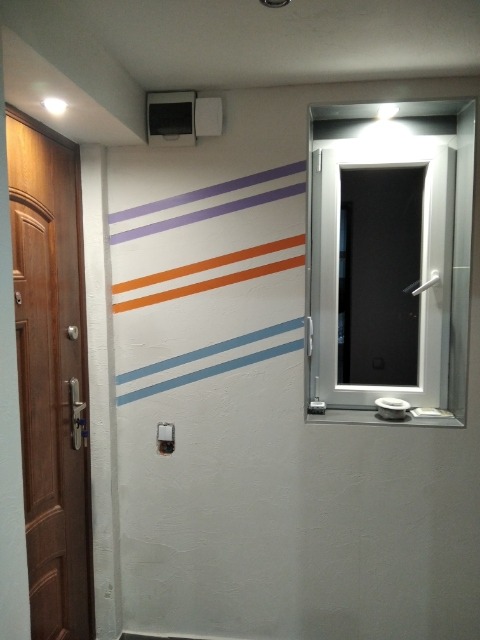 Ściana po malowaniu z poziomymi pasami w kolorach fioletowym, pomarańczowym i niebieskim, obok drewnianych drzwi wejściowych i okna z białymi ramami.