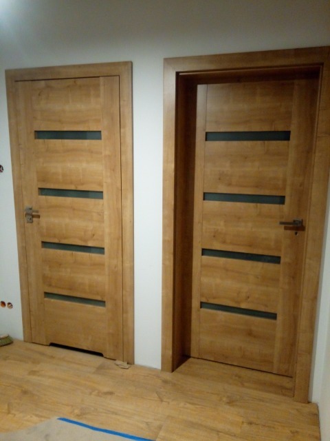 Dwa drewniane, identyczne drzwi wewnętrzne z poziomymi, ciemnymi przeszkleniami, zamontowane w białej ścianie, widoczne z poziomu podłogi imitującej drewno.