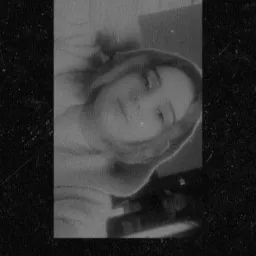 Czarno-białe, ziarniste zdjęcie portretowe młodej kobiety, leżącej na boku, z delikatnym uśmiechem i rozproszonym spojrzeniem, ujęte z góry.