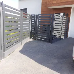 Nowoczesne, szare ogrodzenie panelowe z poziomymi szczeblami i furtką, widoczne z zewnątrz posesji, z fragmentem białego samochodu w prawym dolnym rogu.