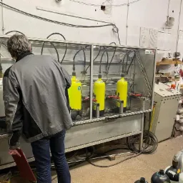 Pracownik kontroluje proces testowania ciśnieniowego butli gazowych w komorze z przezroczystymi ścianami, widoczne manometry i przewody, w tle warsztat z instalacjami na ścianie.