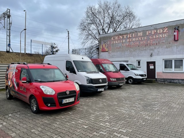 Flota pojazdów firmy z Wrocławia specjalizującej się w usługach przeciwpożarowych, zaparkowana przed budynkiem z napisem 'Centrum PPOŻ Zakład Remontu Gaśnic'.