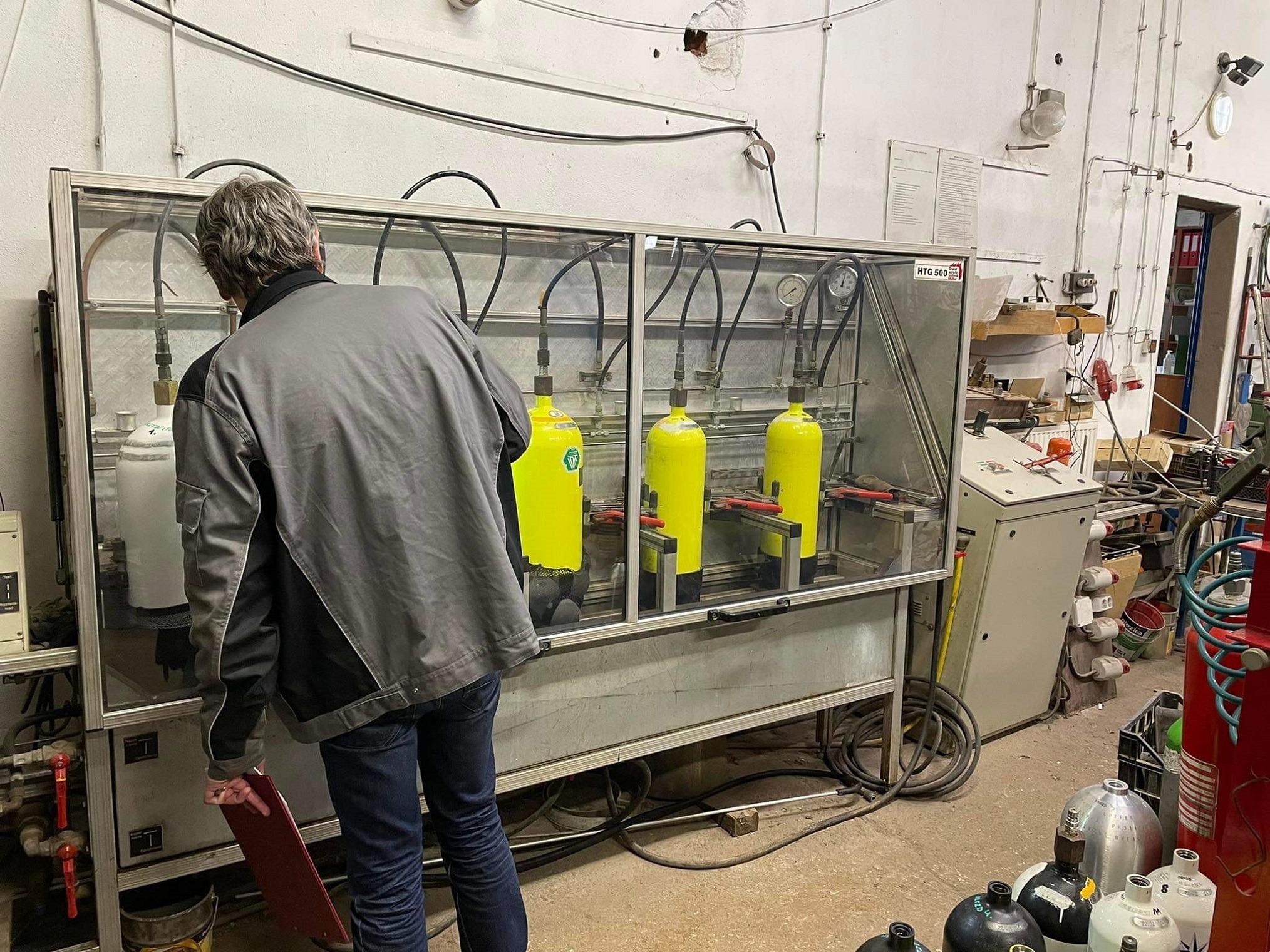 Pracownik kontroluje proces testowania ciśnieniowego butli gazowych w komorze z przezroczystymi ścianami, widoczne manometry i przewody, w tle warsztat z instalacjami na ścianie.