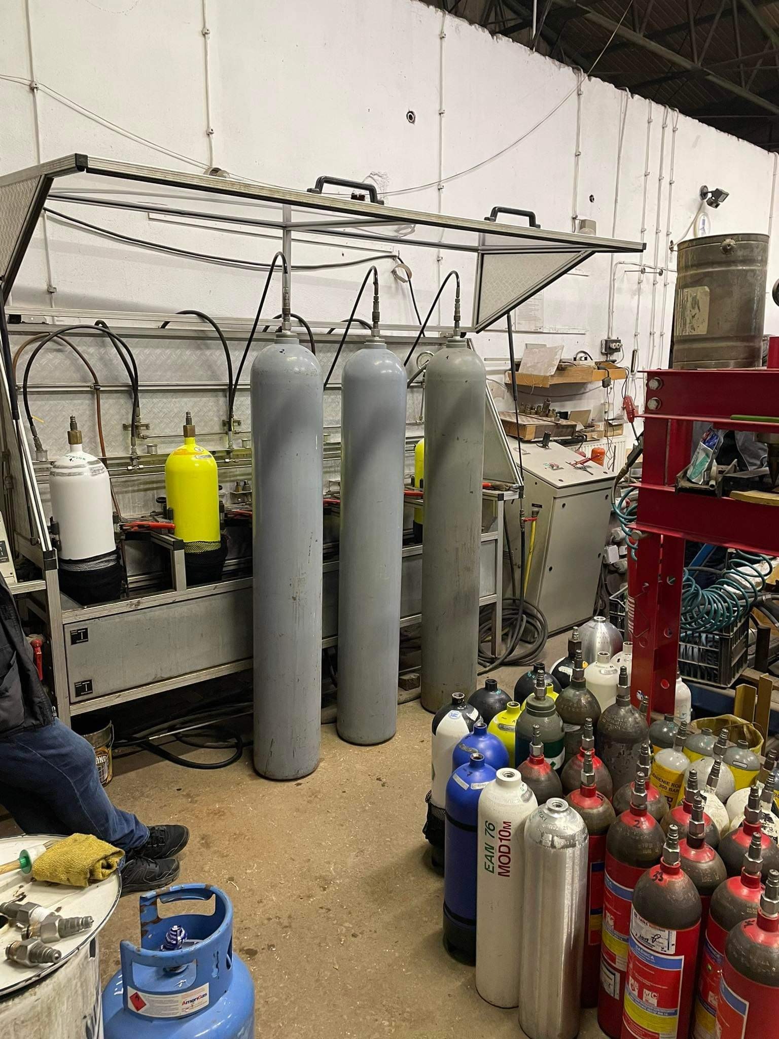 Wnętrze warsztatu z urządzeniem do testowania butli ciśnieniowych, szereg szarych butli podłączonych do urządzenia, oraz zgromadzone na podłodze butle w różnych kolorach i rozmiarach.