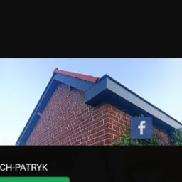 Fragment dachu z czerwonej dachówki i ciemnoszarej obróbki blacharskiej, widok z dołu na tle błękitnego nieba, ściana budynku z czerwonej cegły.