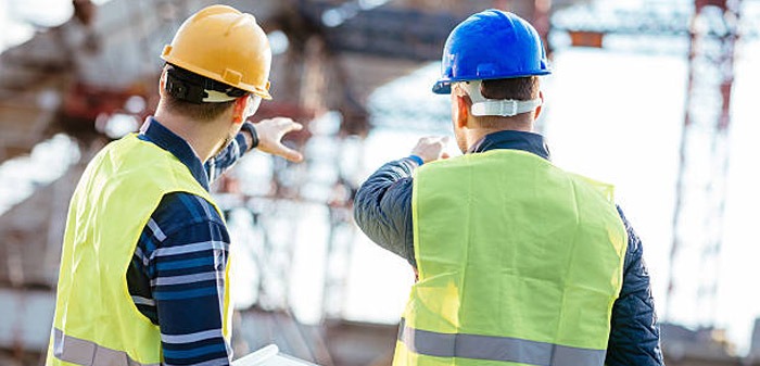 Dwóch inżynierów w kaskach (żółtym i niebieskim) i kamizelkach odblaskowych omawia projekt konstrukcji stalowej na placu budowy, jeden z nich wskazuje palcem na elementy konstrukcyjne.