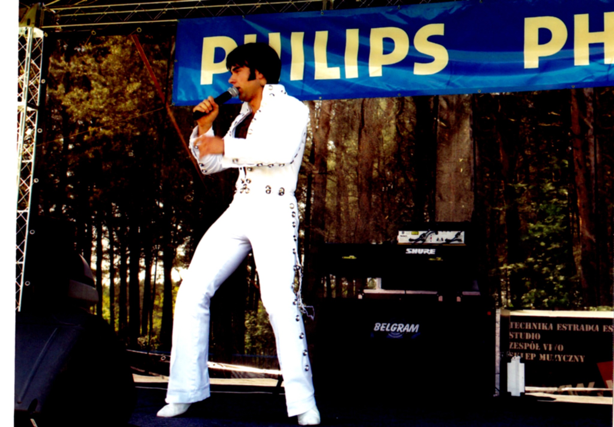 Mężczyzna w białym kostiumie w stylu Elvisa Presleya śpiewa do mikrofonu na scenie, w tle las i baner z napisem.