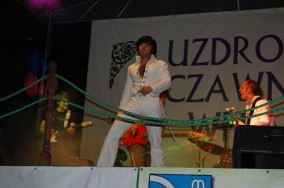 Wokalista w białym kostiumie w stylu Elvisa Presleya, występuje na scenie z zespołem podczas wydarzenia plenerowego, z widocznym banerem w tle i zabezpieczeniem linowym przed sceną.