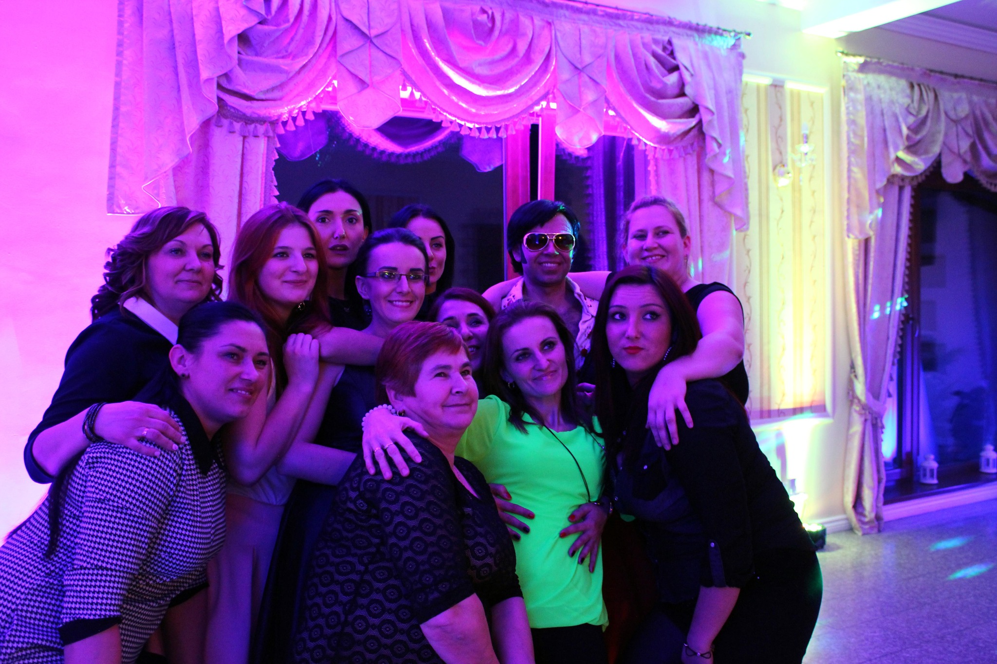 Grupa kobiet i mężczyzna w okularach pozuje do zdjęcia na tle dekoracji okiennej w sali oświetlonej kolorowym światłem.