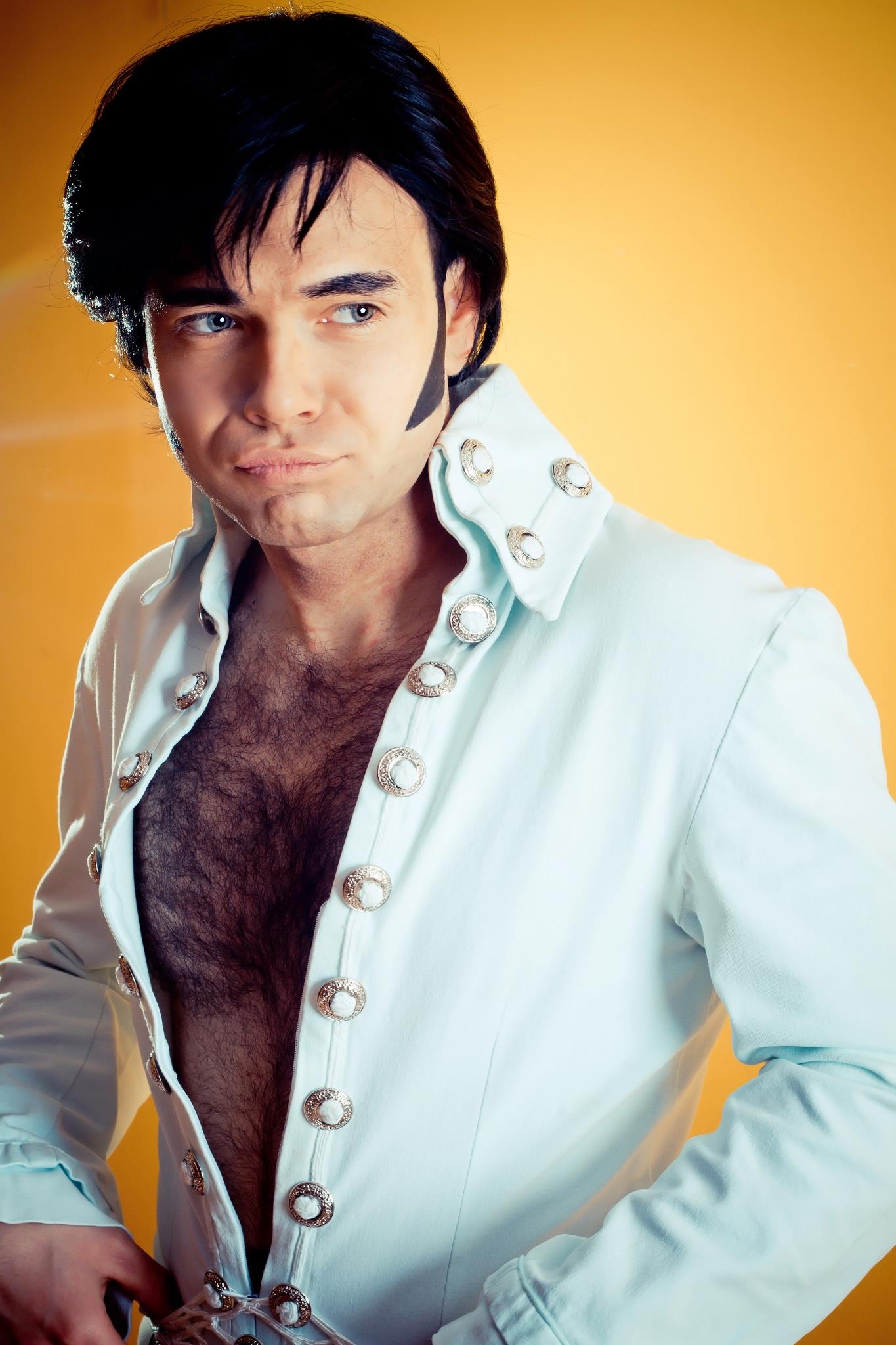 Mężczyzna w kostiumie scenicznym w stylu Elvisa Presleya, z charakterystyczną fryzurą i bokobrodami, ubrany w rozpiętą koszulę z ozdobnymi guzikami, na jednolitym żółtym tle.