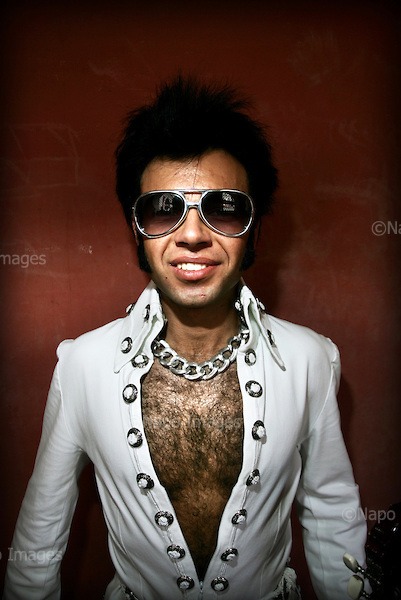 Wokalista z Poznania w kostiumie stylizowanym na Elvisa Presleya, z charakterystyczną fryzurą i okularami przeciwsłonecznymi, pozuje na tle czerwonej ściany.