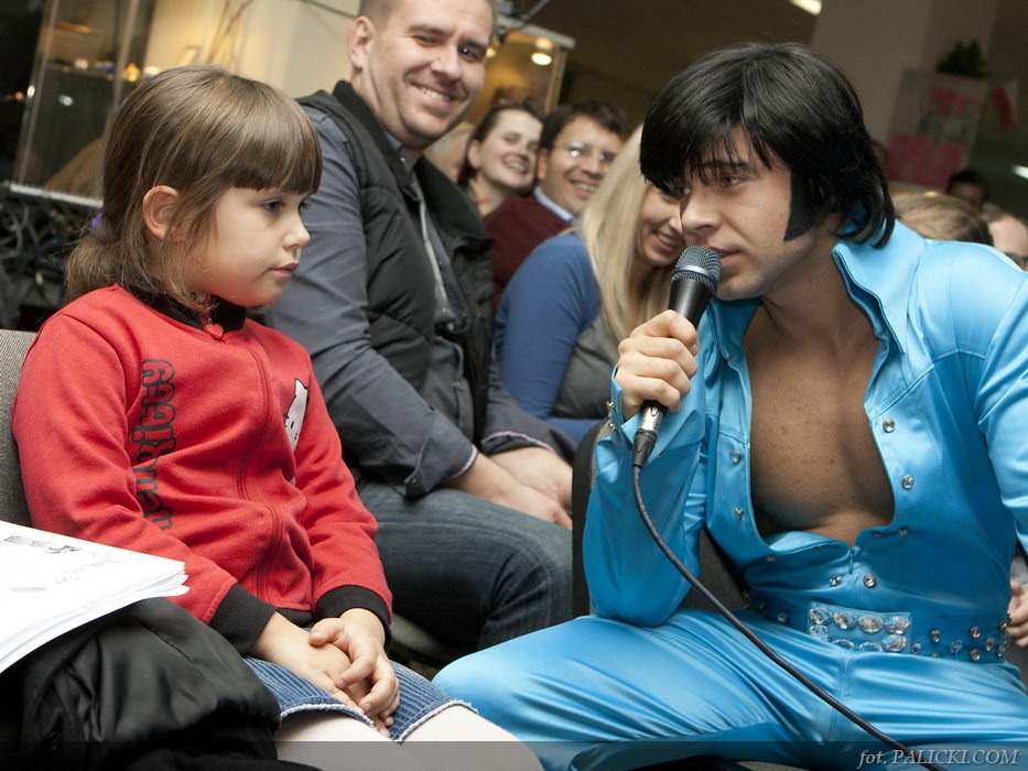 Występujący mężczyzna przebrany za Elvisa Presleya z mikrofonem, w niebieskim kombinezonie, śpiewa dla publiczności, w której siedzi mała dziewczynka w czerwonej bluzie.