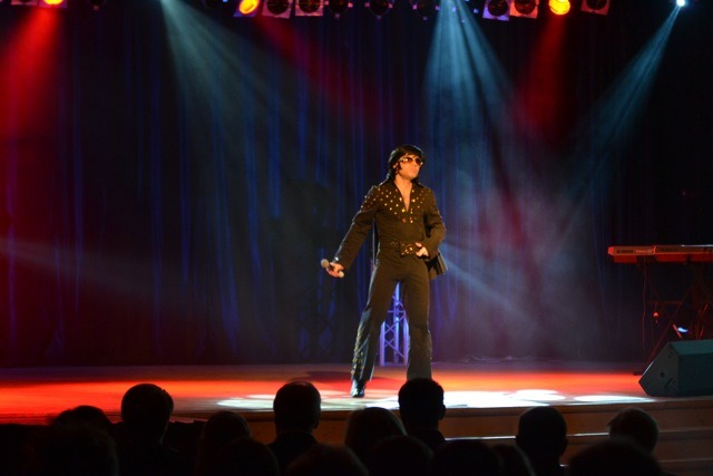 Mężczyzna przebrany za Elvisa Presleya występuje na scenie w kostiumie z cekinami, trzyma mikrofon i ma ciemne okulary; w tle widoczna publiczność, reflektory i instrumenty muzyczne.