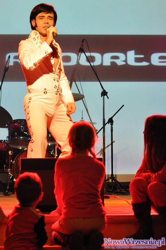 Młody mężczyzna przebrany za Elvisa Presleya śpiewa do mikrofonu na scenie podczas występu dla dzieci, w tle widoczny perkusja i logo Oferteo.