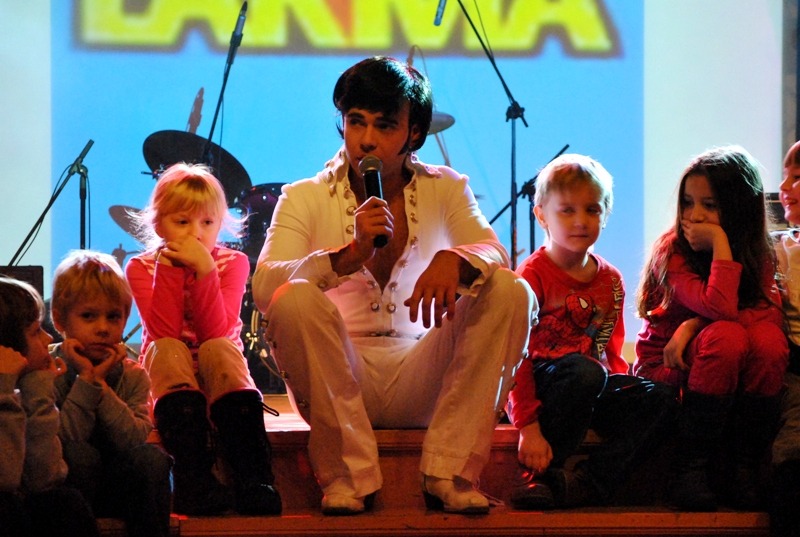Mężczyzna przebrany za Elvisa Presleya śpiewa do mikrofonu, siedząc na scenie otoczony grupą zaciekawionych dzieci.