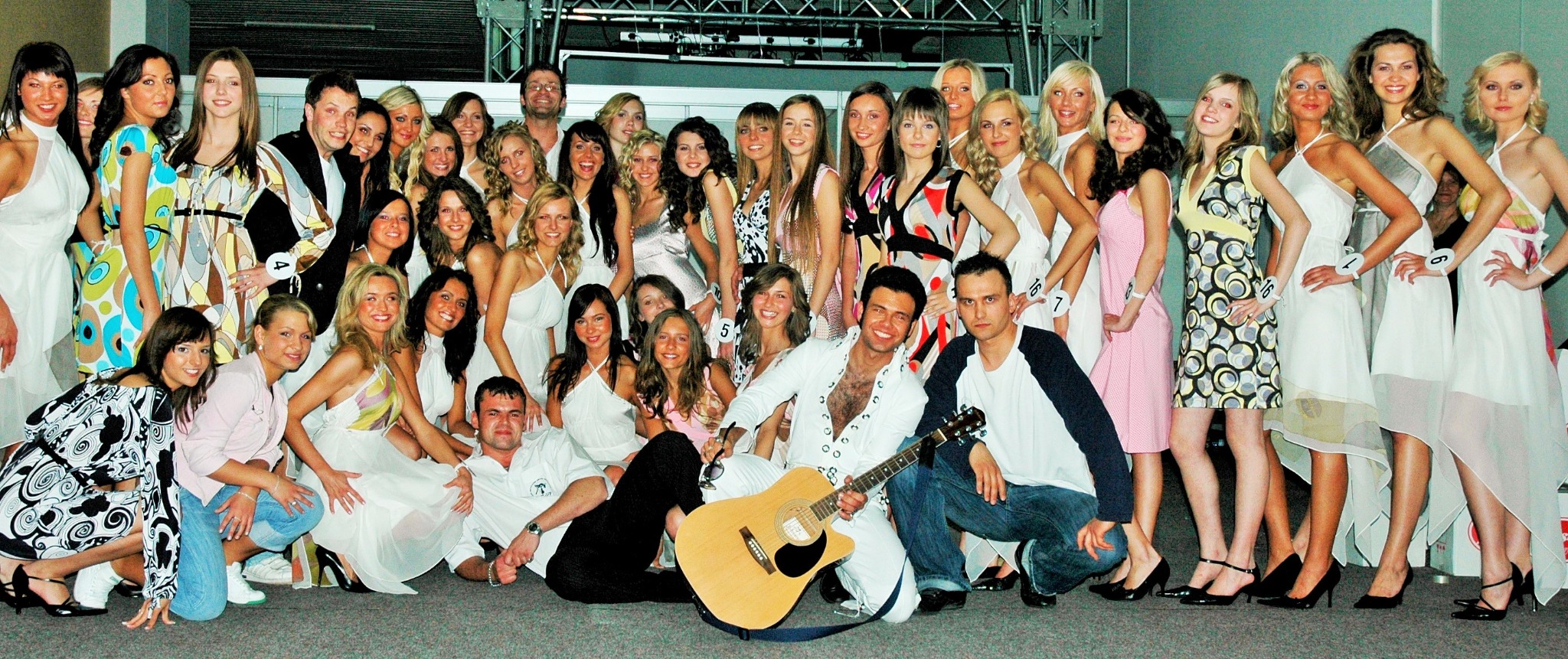 Duża grupa kobiet w sukienkach, prawdopodobnie uczestniczek konkursu piękności, pozuje do zdjęcia z dwoma mężczyznami, jeden w stroju imitującym Elvisa Presleya z gitarą akustyczną.