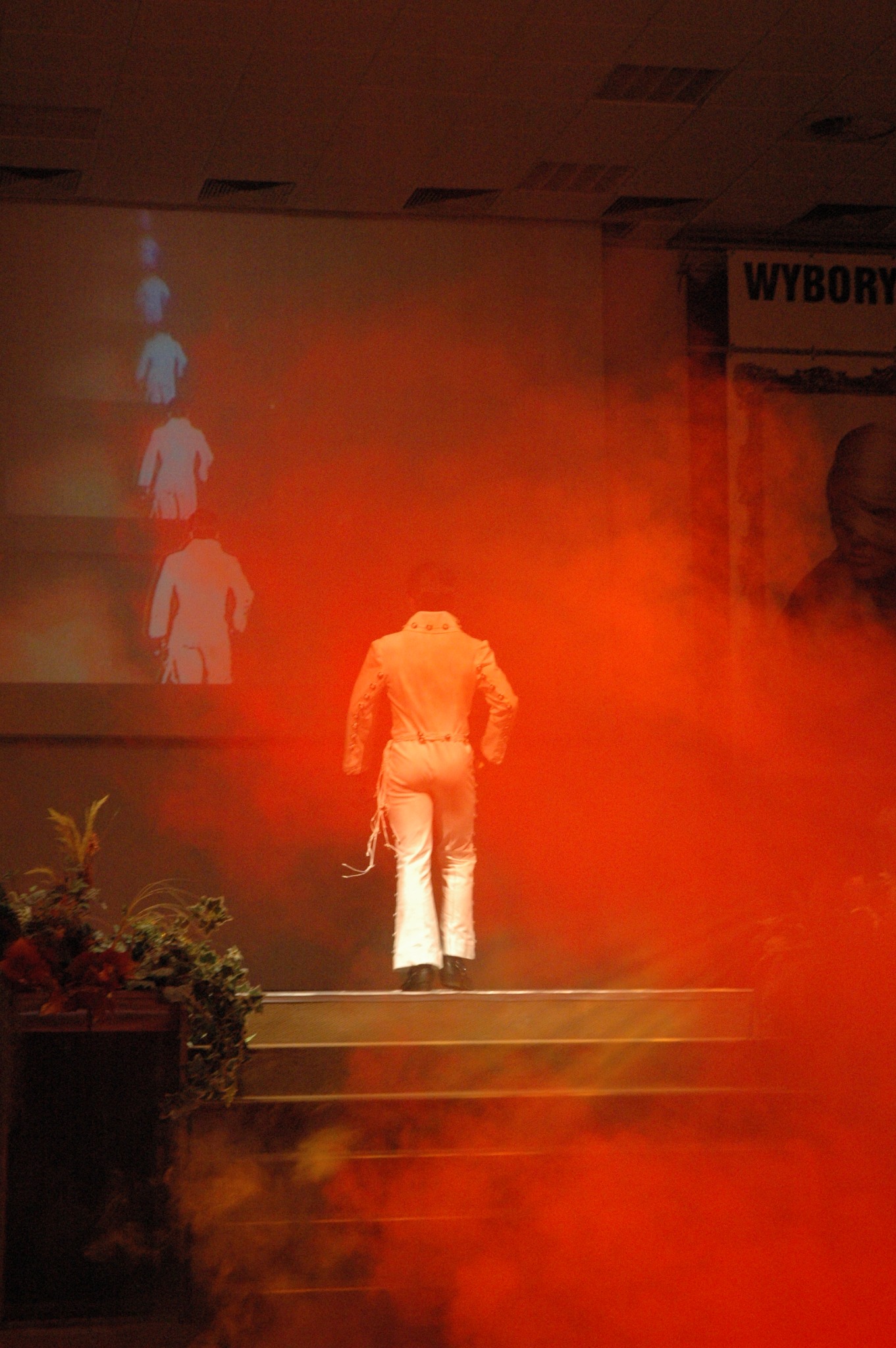 Artysta w białym kombinezonie wchodzi po schodach na scenę w czerwonym oświetleniu, z projekcją sylwetek w tle i dymem scenicznym.