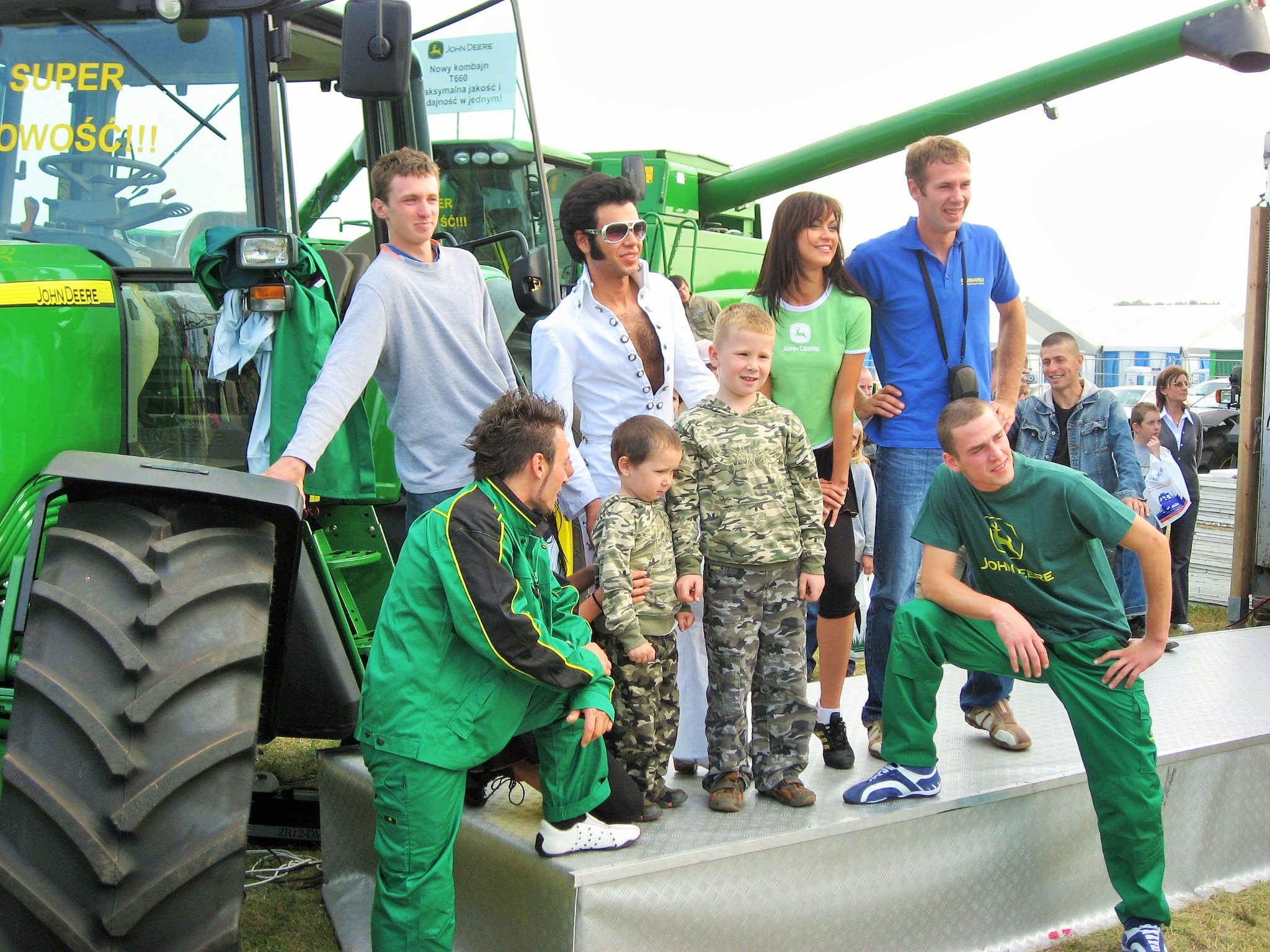 Grupa osób pozuje na tle nowoczesnego, zielonego kombajnu John Deere na plenerowej prezentacji. Wśród nich mężczyzna przebrany za Elvisa Presleya.