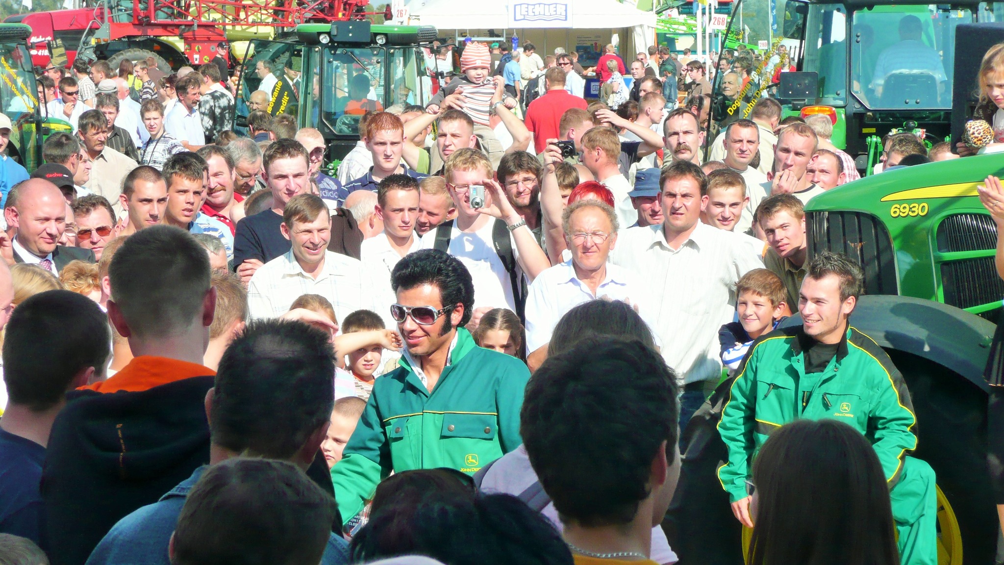 Tłum ludzi na otwartej przestrzeni, w tle traktory i budki wystawiennicze, w pierwszym planie mężczyzna przebrany za Elvisa w zielonej kurtce z logo John Deere.