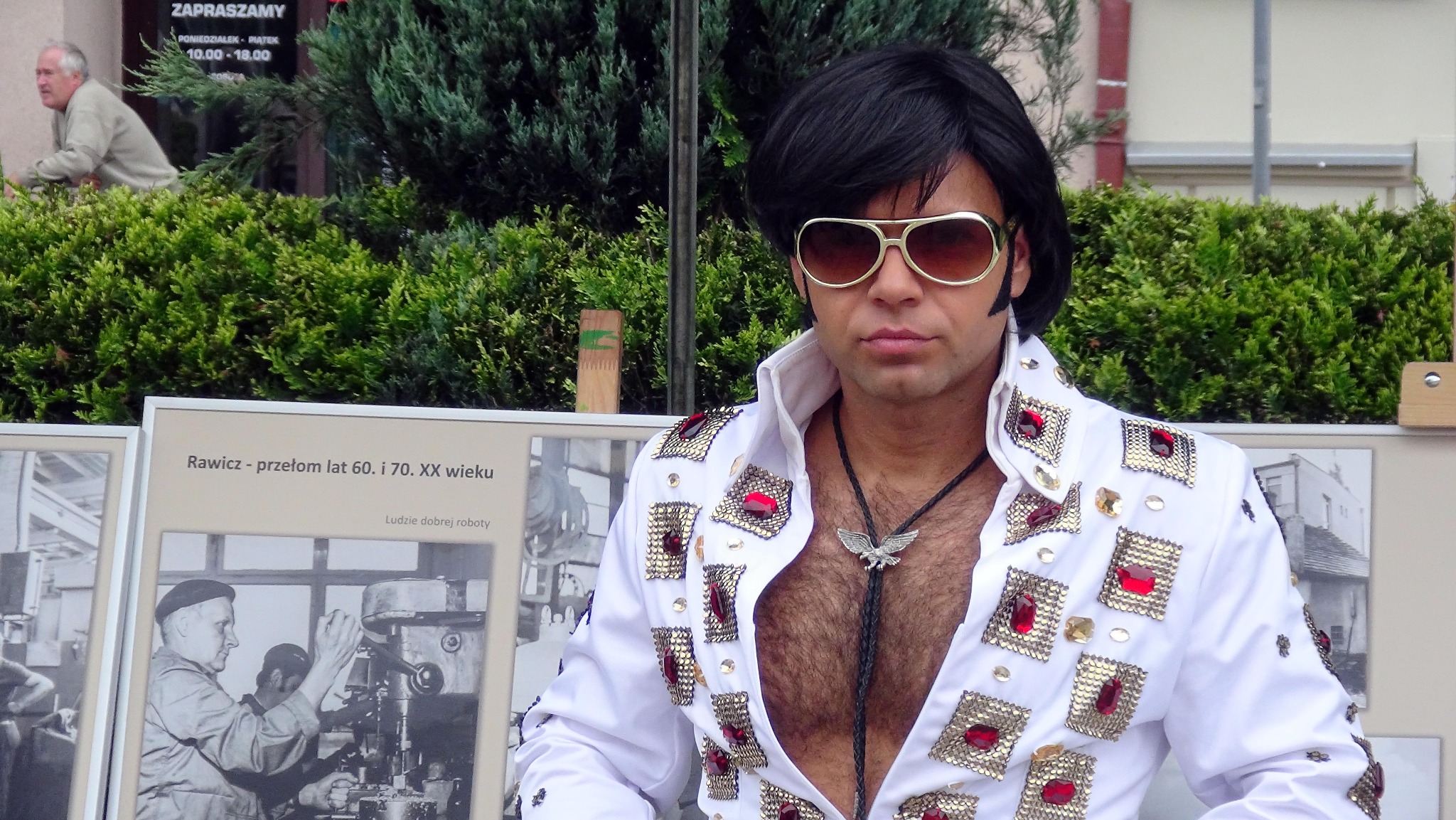 Mężczyzna przebrany za Elvisa Presleya w białym kostiumie z czerwonymi i złotymi zdobieniami, pozujący na tle zielonych krzewów i wystawy fotograficznej.