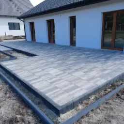 Nowo ułożony taras z szarej kostki brukowej z ciemnymi obrzeżami przy białym domu z brązowymi oknami, widoczne przygotowane podłoże i częściowo ułożone obrzeża.
