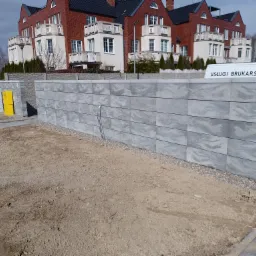 Nowo wzniesiony, szary mur z betonowych bloczków o falistym wzorze, widoczny na tle budynku mieszkalnego i zaparkowanych samochodów, z zaparkowanym vanem z napisem 'Usługi Brukarskie'.