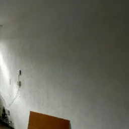 Ściana po nałożeniu gładzi strukturalnej, widoczne gniazdka elektryczne, fragment okna z widokiem na rusztowanie i resztki materiałów budowlanych na podłodze.