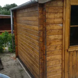 Drewniany domek ogrodowy z brązowych desek, widoczny róg budynku z drzwiami, szara rynna i fragment niebieskiego krzesła na pierwszym planie.