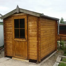 Drewniana szopa ogrodowa z małym oknem i spadzistym dachem, obok szklarni i zarośniętego ogrodu.