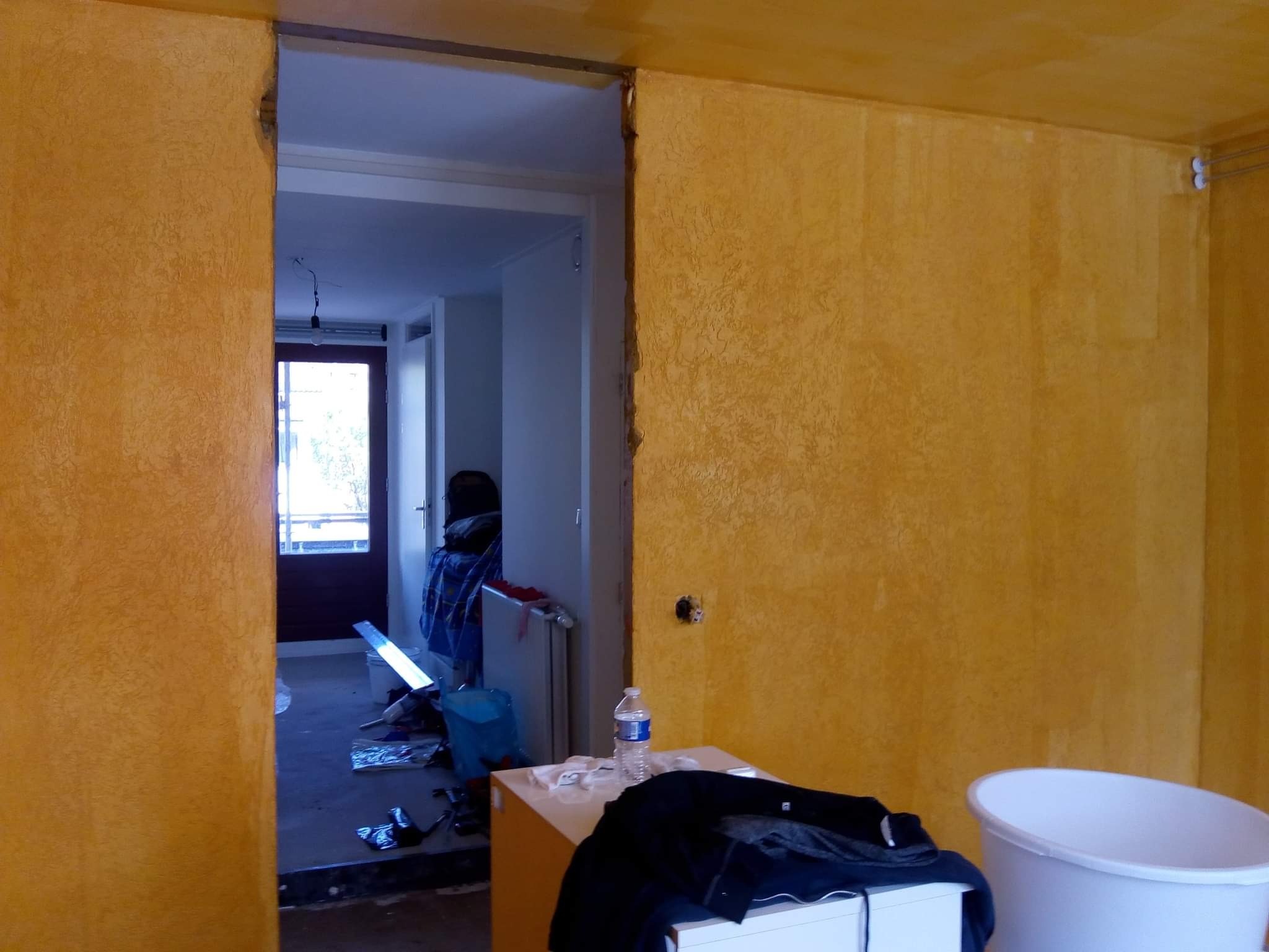 Pokój w trakcie remontu: żółte ściany z widoczną fakturą, uszkodzone obramowanie drzwi, fragment widocznego wnętrza z pozostawionymi narzędziami i materiałami budowlanymi, butelka wody i ubranie...