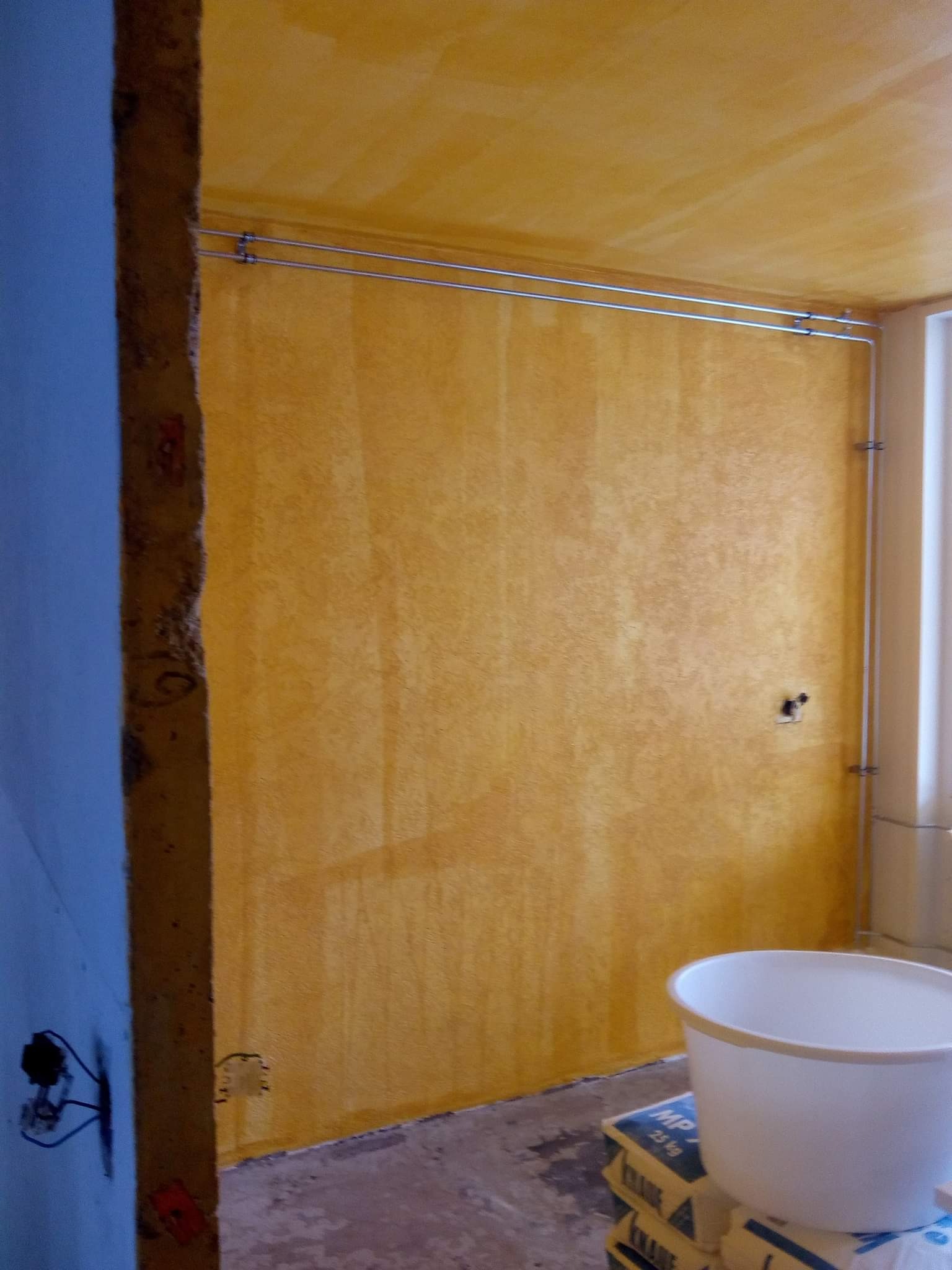 Żółta ściana po nałożeniu gładzi, widoczne ślady po wałku, instalacja elektryczna na ścianie, wiadro i worki z gładzią Knauf MP 75 na podłodze.