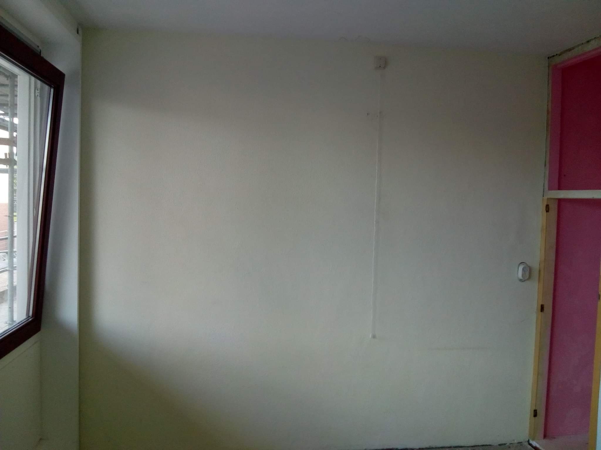 Wnętrze pokoju po malowaniu ścian na jasny odcień, widoczny fragment okna z brązową ramą, ściana z wystającym kablem i gniazdkiem oraz fragment futryny pomalowanej na różowo