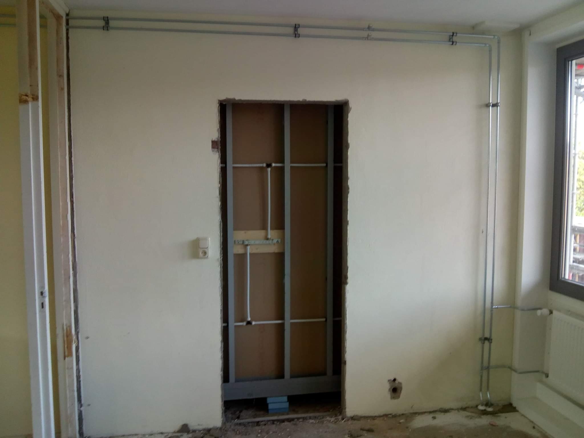 Przygotowanie do montażu ościeżnicy: metalowa konstrukcja szkieletowa w otworze drzwiowym, widoczne instalacje elektryczne na ścianie, fragment drewnianej ramy po lewej stronie.