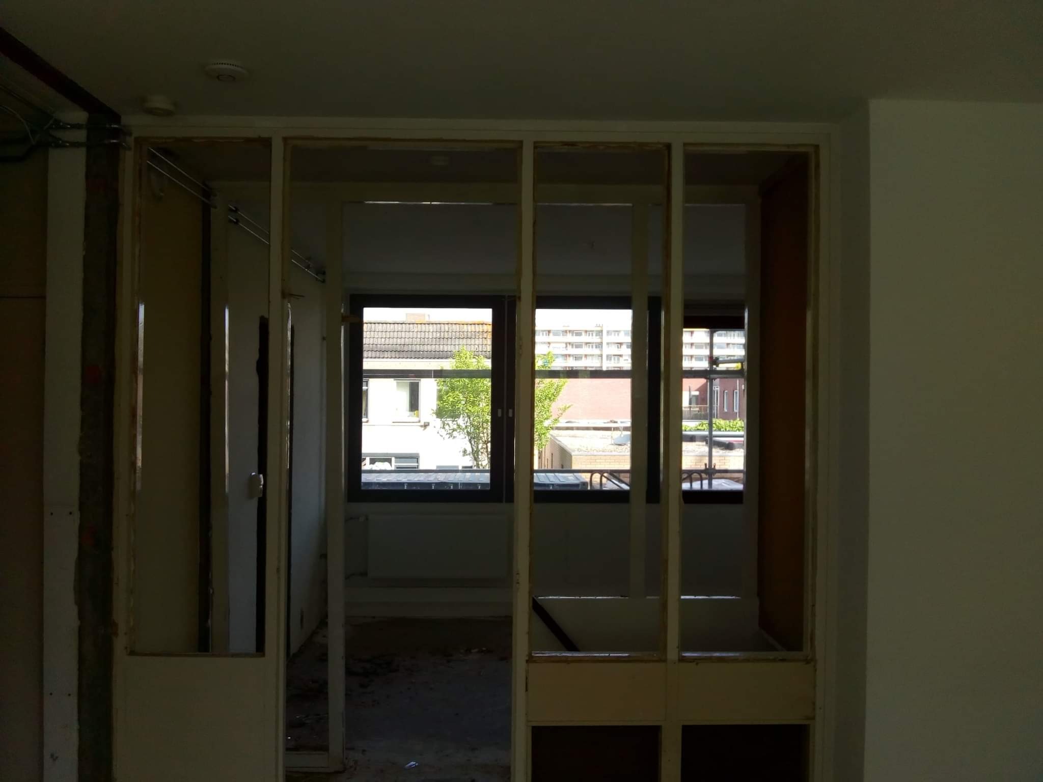 Demontaż ściany działowej w mieszkaniu, widoczne odsłonięte elementy konstrukcyjne, okna z widokiem na budynki i zieleń.