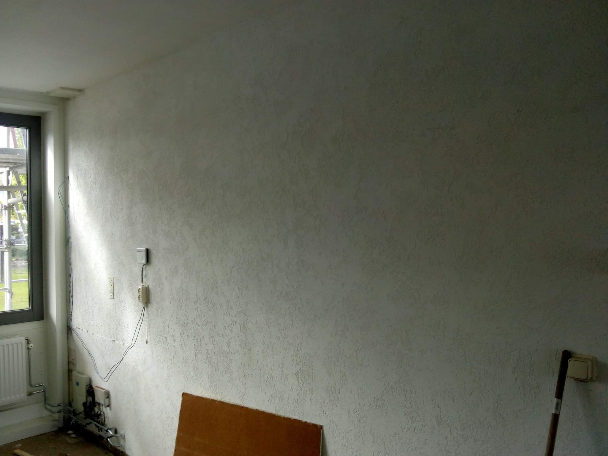 Ściana po nałożeniu gładzi strukturalnej, widoczne gniazdka elektryczne, fragment okna z widokiem na rusztowanie i resztki materiałów budowlanych na podłodze.