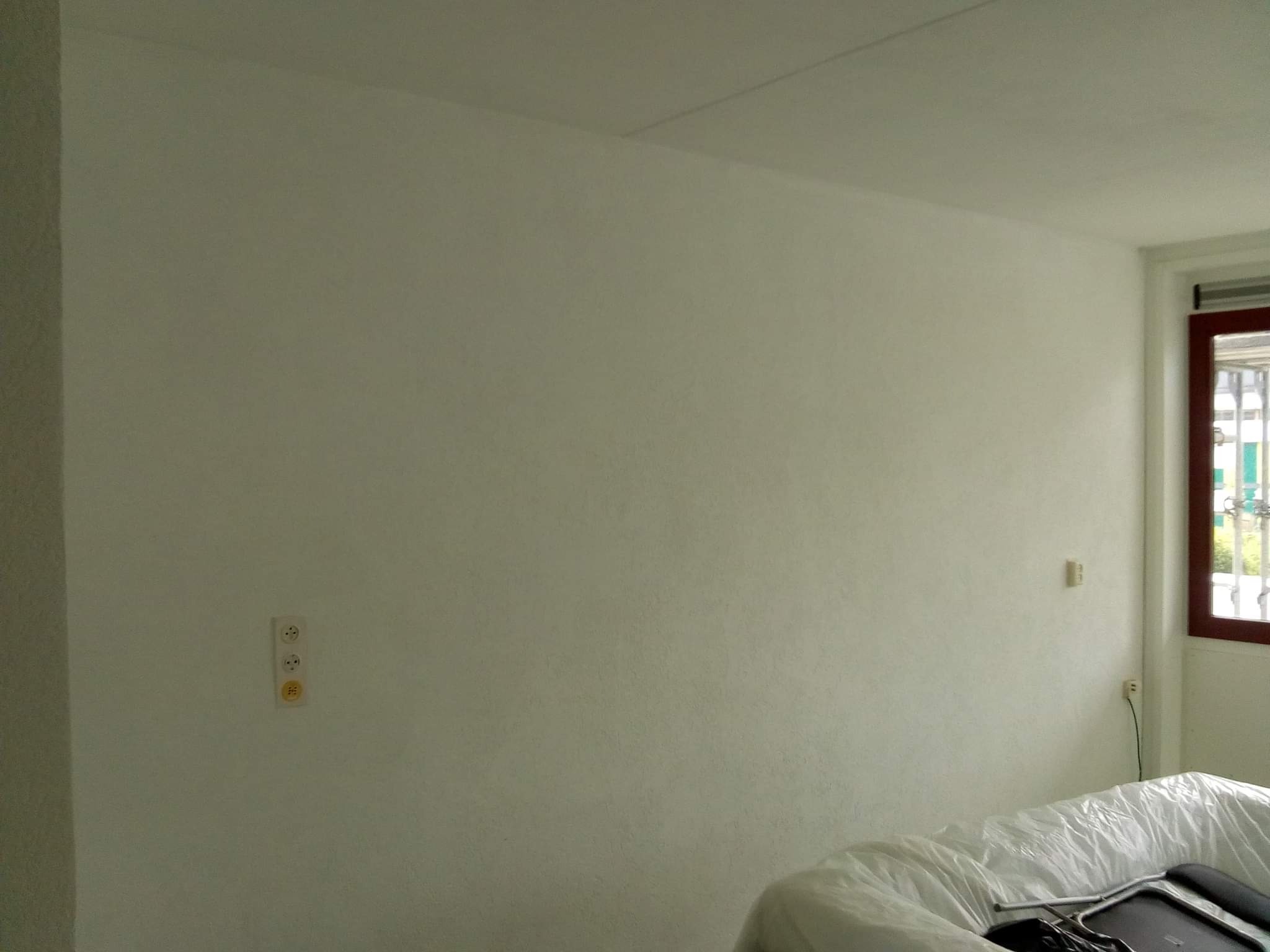 Świeżo pomalowana na biało ściana z widocznymi gniazdkami elektrycznymi, zabezpieczone meble folią malarską w dolnej części kadru.