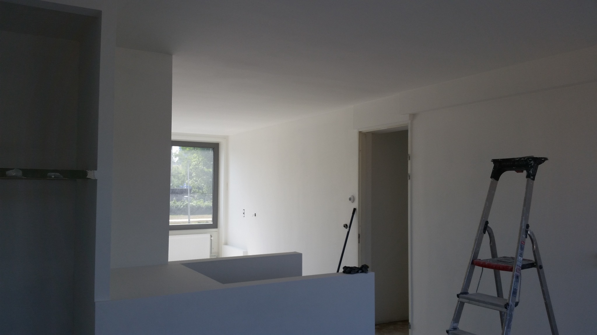 Wnętrze świeżo pomalowanego na biało pomieszczenia z widoczną drabiną malarską, fragmentem zabudowy i oknem z widokiem na zieleń.