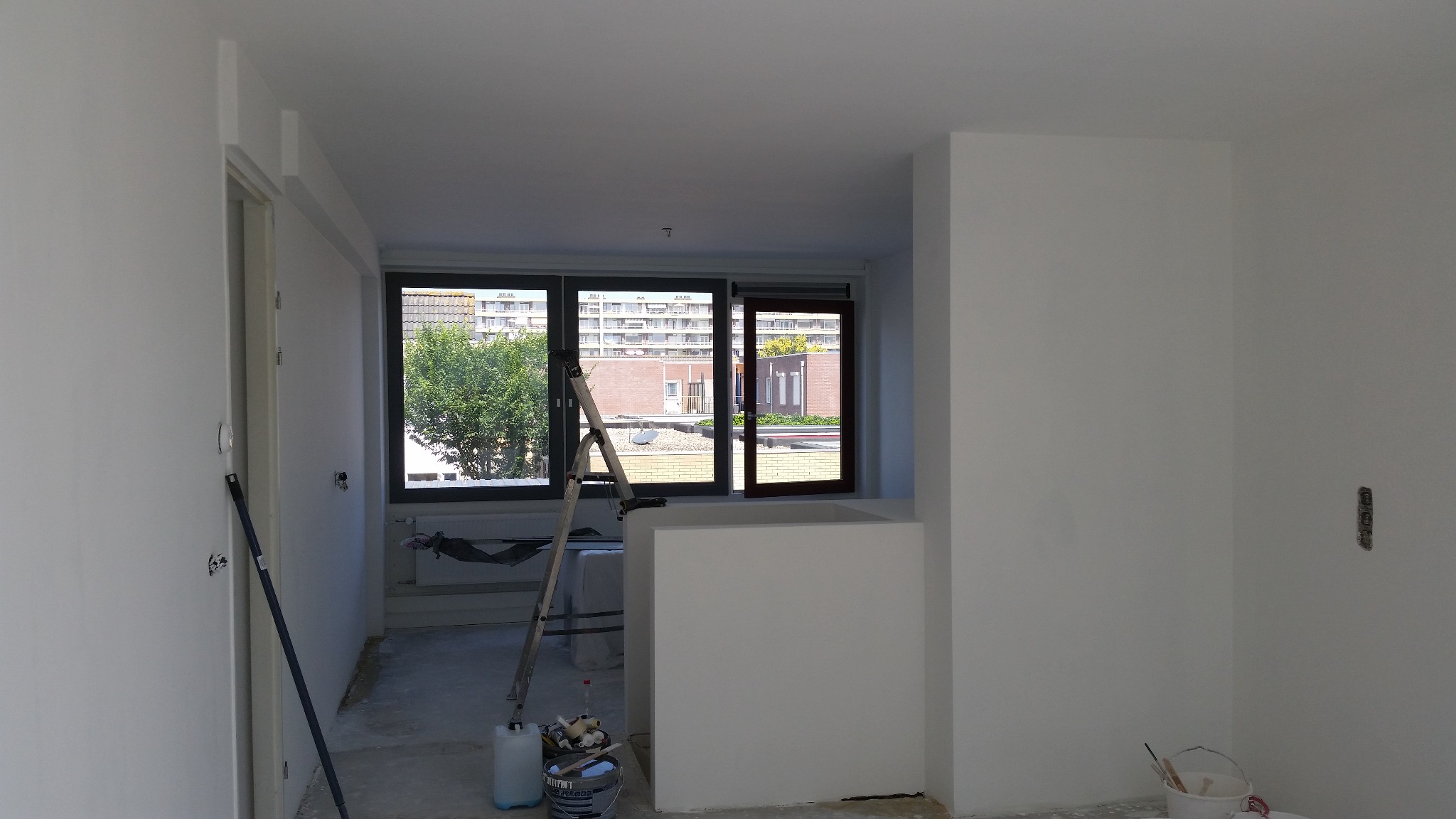 Wnętrze pokoju w trakcie odświeżania, z widoczną drabiną opartą o okno, pojemnikami z farbą i świeżo pomalowanymi na biało ścianami. Widoczne niedoskonałości w malowaniu przy podłodze.