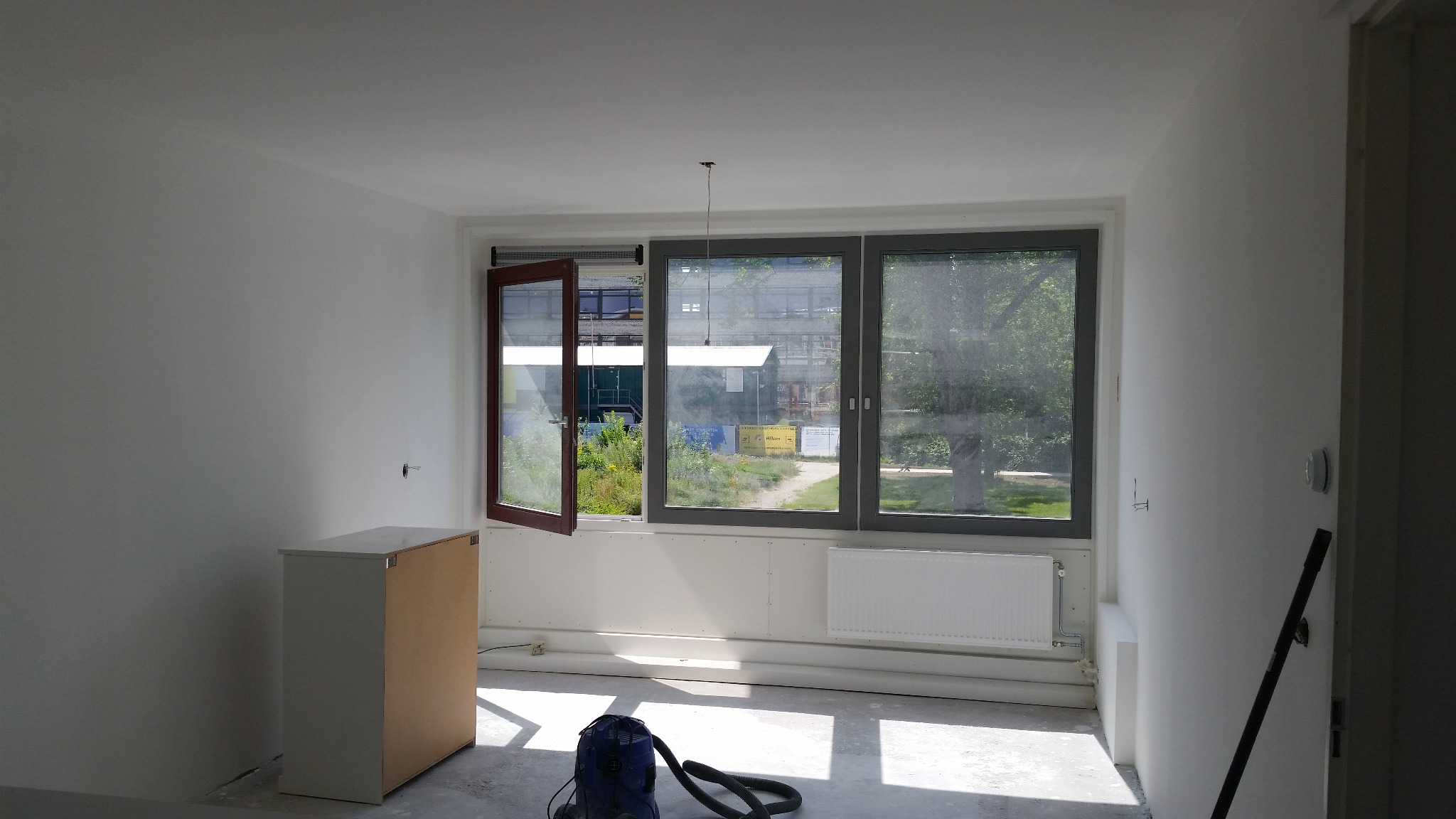 Wnętrze pokoju po malowaniu ścian na biało, widoczne okna z widokiem na zieleń, grzejnik, szafka, odkurzacz i przewód elektryczny wystający z sufitu.