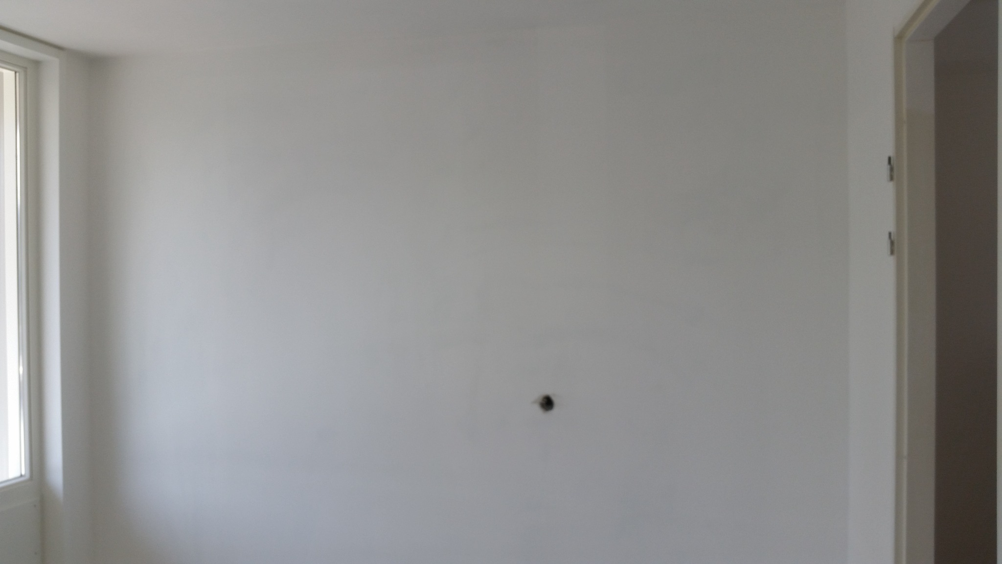 Ściana po nałożeniu gładzi z widocznym otworem na gniazdko elektryczne i fragmentem okna po lewej stronie.