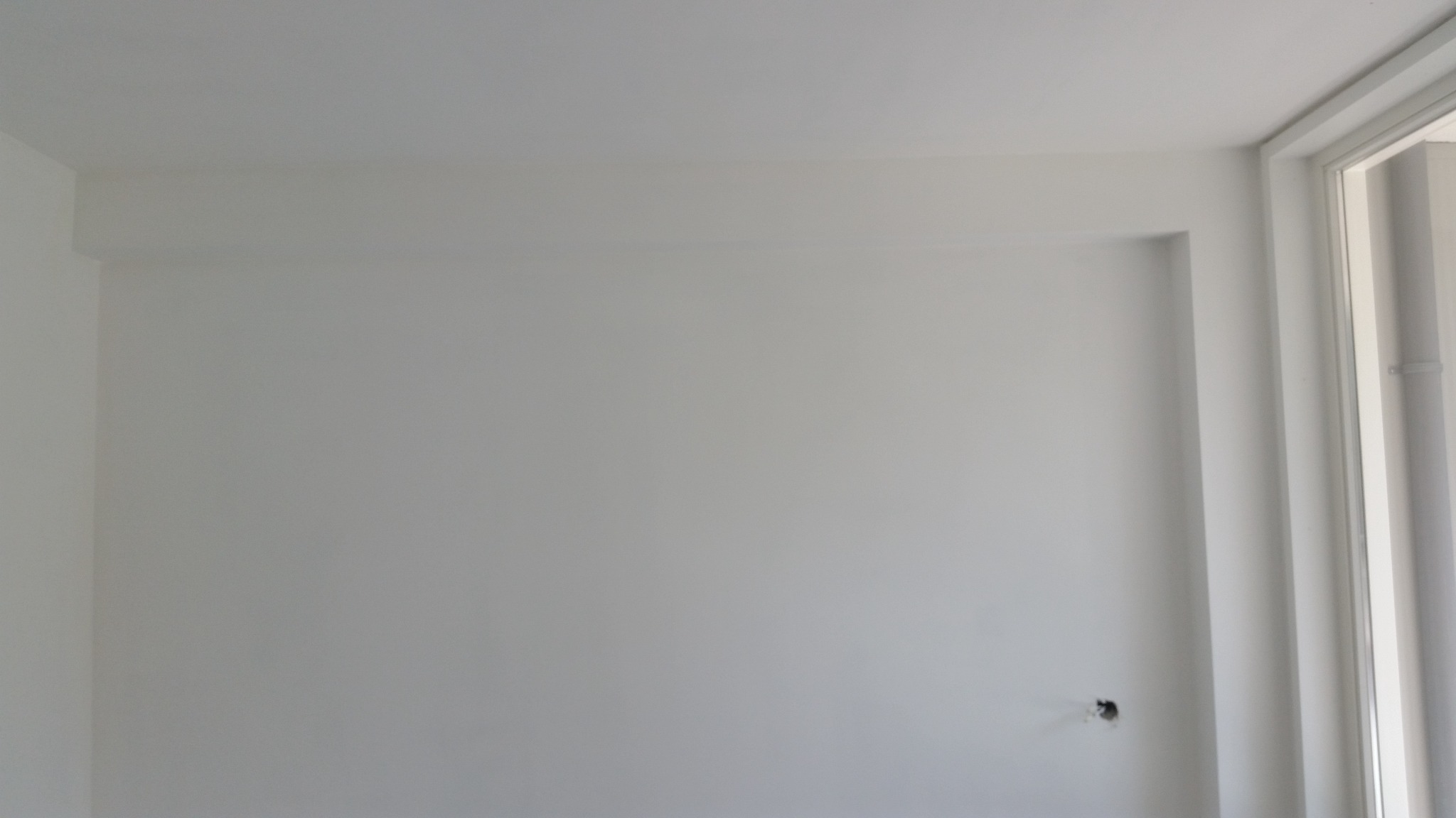 Wnętrze pokoju po malowaniu na biało, widoczna świeżo pomalowana ściana z gładką powierzchnią, fragment okna z białymi ramami po prawej stronie i otwór po gniazdku elektrycznym w dolnej części ściany.