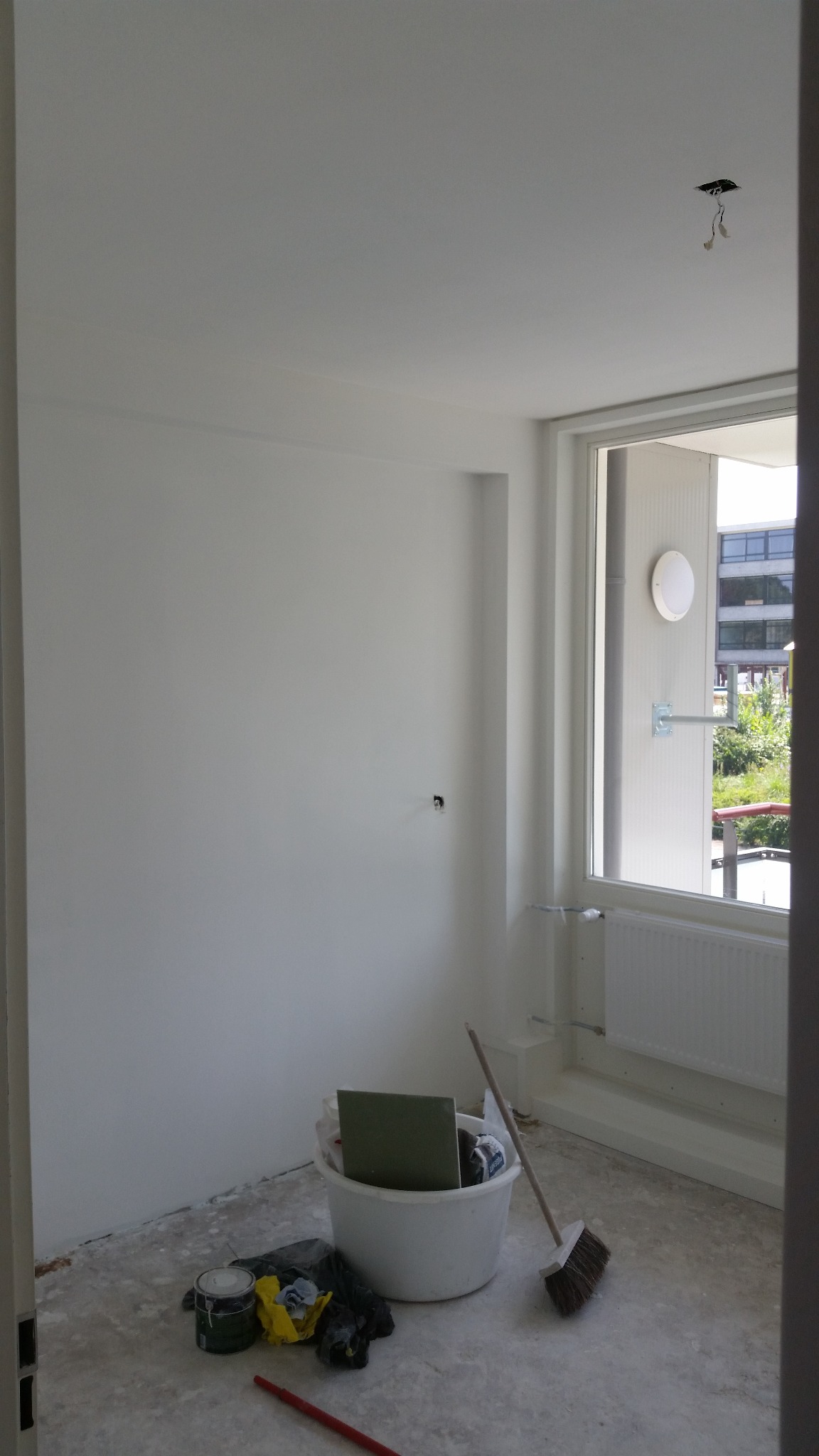 Wnętrze pokoju w trakcie remontu: świeżo pomalowane na biało ściany i sufit, widoczne narzędzia malarskie, pędzel oparty o ścianę, wiadro z akcesoriami, odkryte kable oświetleniowe w suficie, okno...