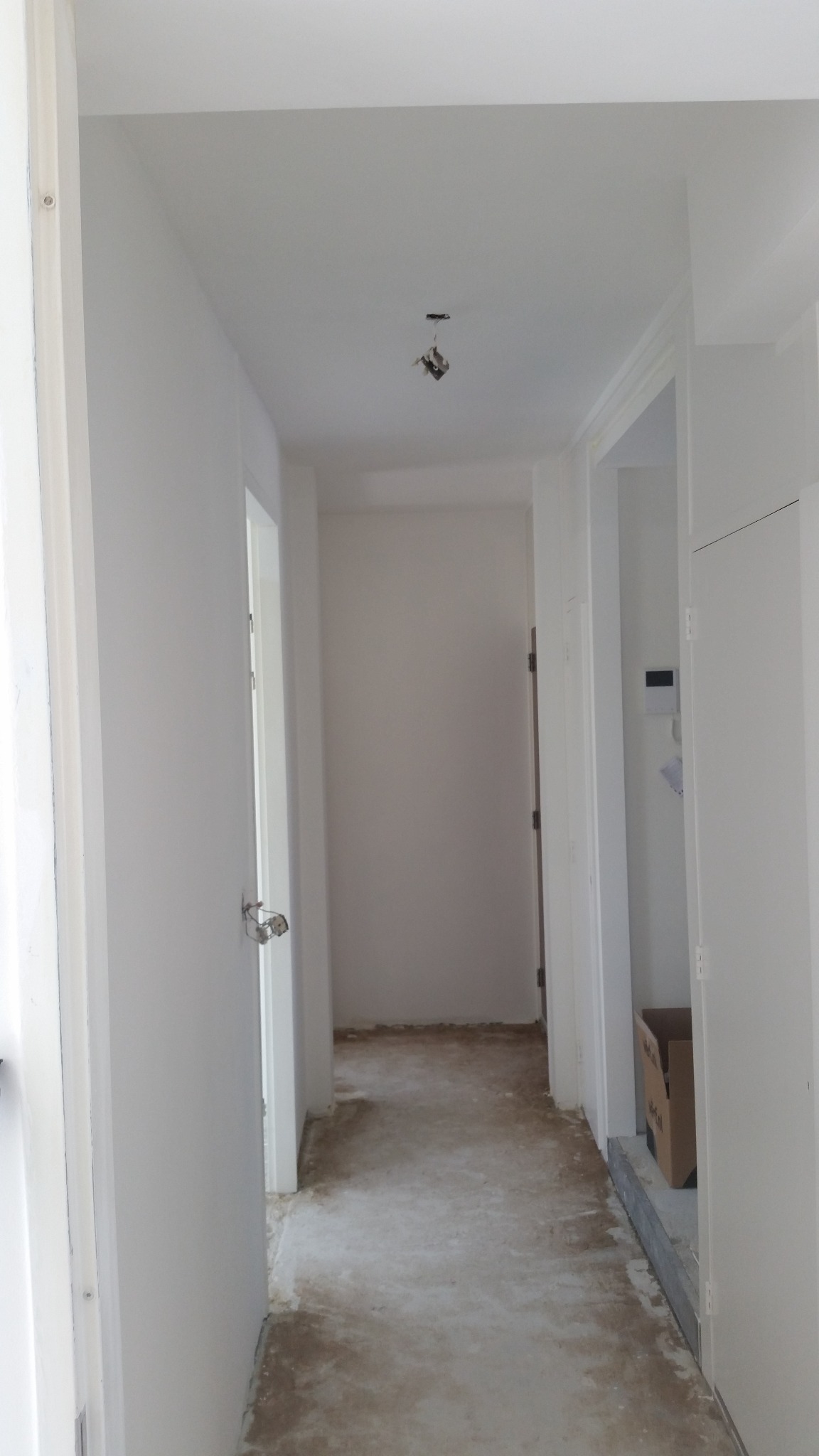 Korytarz w trakcie remontu, widoczne ściany po malowaniu na biało, podłoga przygotowana do dalszych prac, wystające przewody z sufitu, otwarte drzwi po lewej stronie.