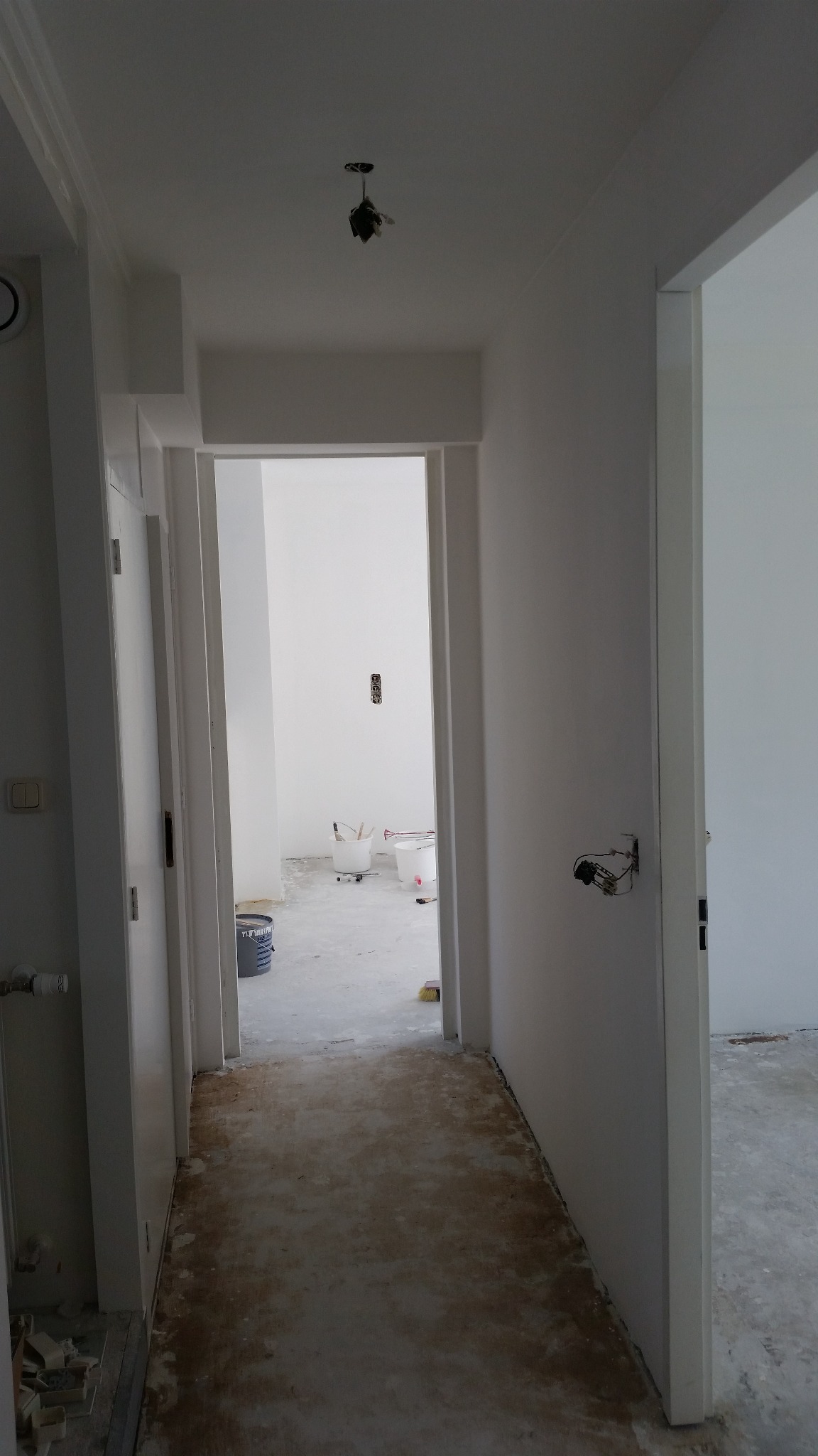 Korytarz w trakcie odświeżania: świeżo pomalowane białe ściany, widoczne ślady po pracach na podłodze, puszki z farbą w tle, niedokończone podłączenie oświetlenia sufitowego.