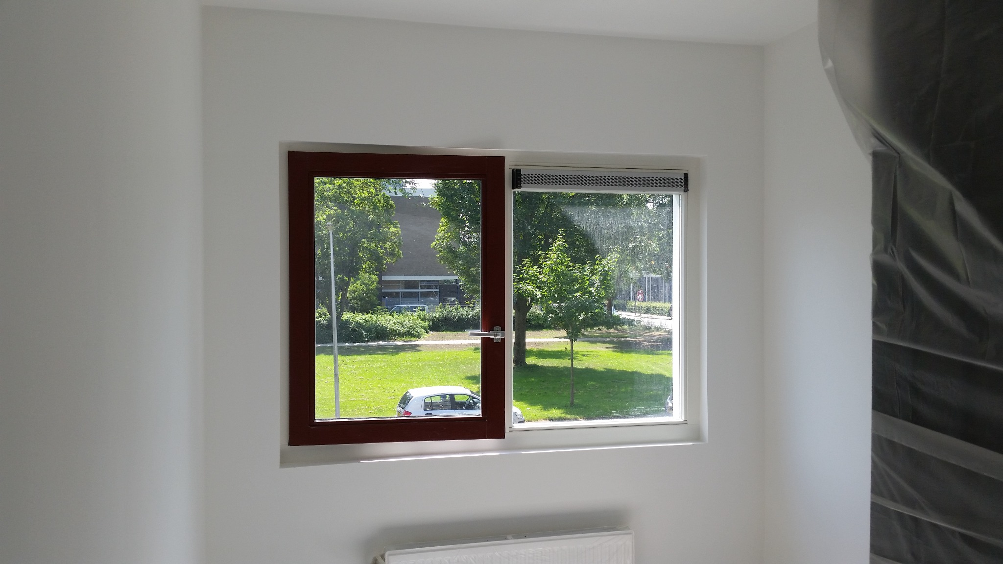 Białe ściany po świeżym malowaniu, widoczne okno z brązową ramą i widokiem na zieleń, grzejnik pod oknem zabezpieczony folią malarską, narożnik pokoju z folią ochronną.