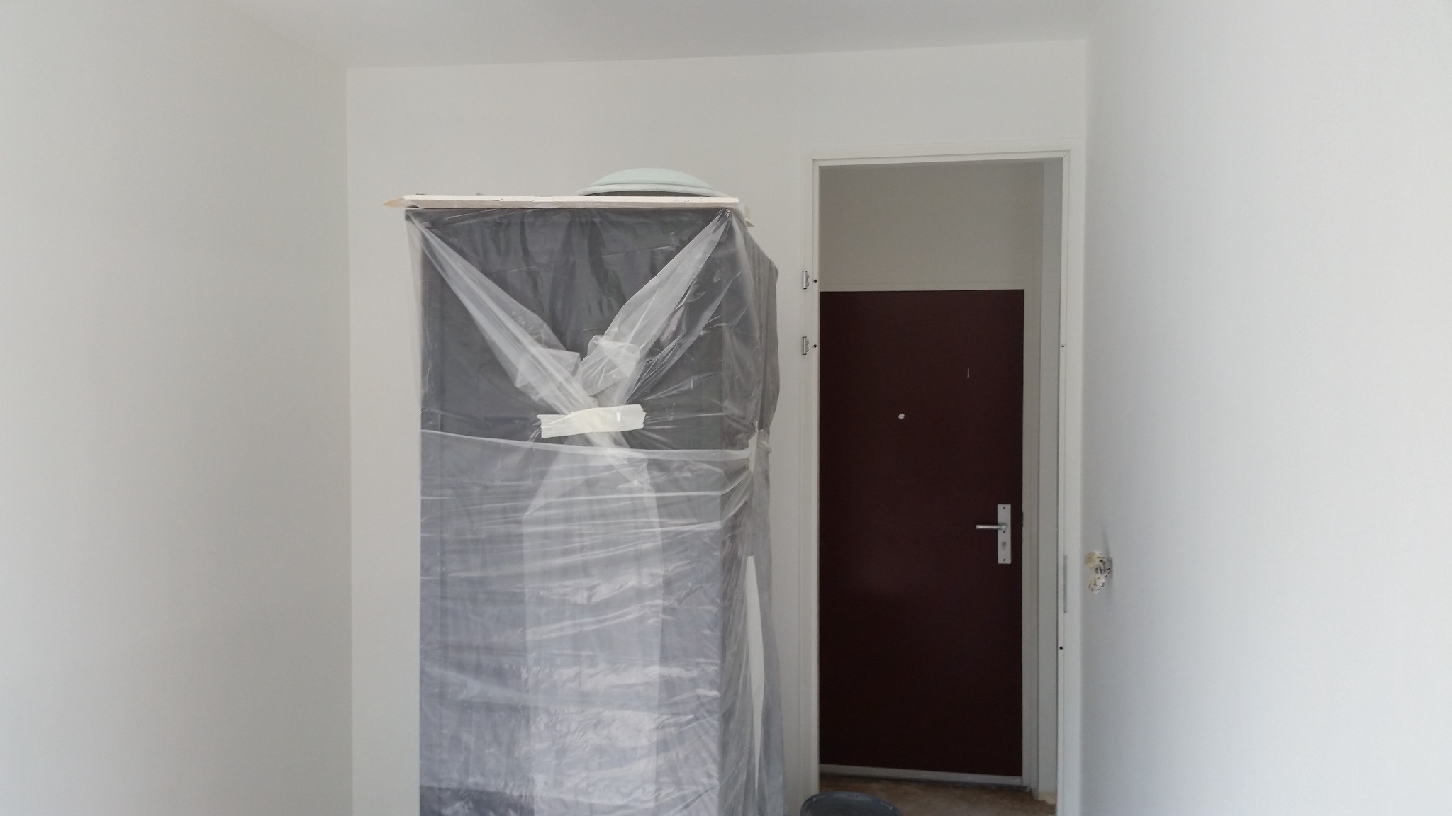 Przygotowanie do malowania: szafa zabezpieczona folią malarską w nowo wykończonym, jasnym pomieszczeniu z widoczną częścią brązowych drzwi.