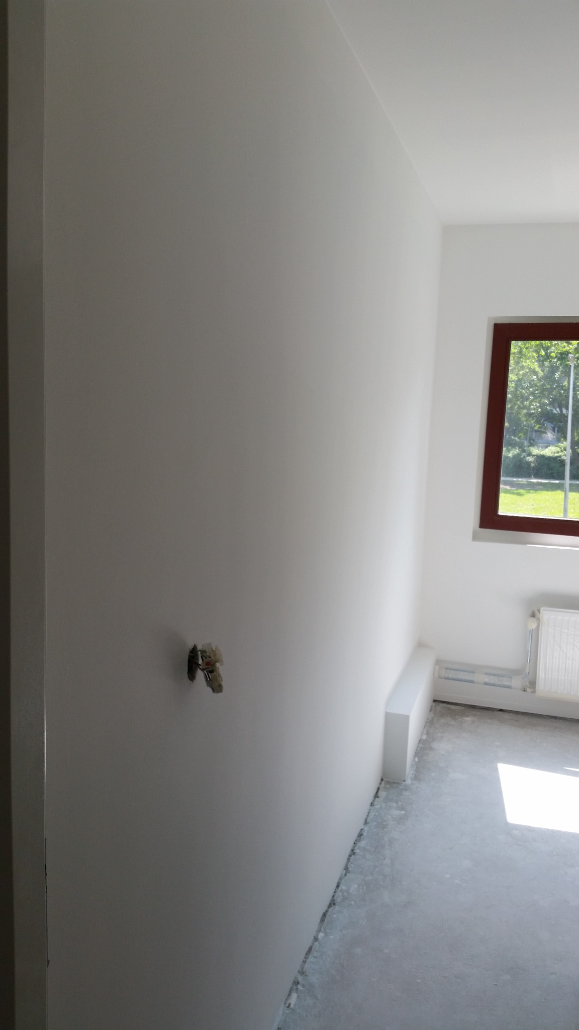 Świeżo pomalowane na biało wnętrze pokoju z widocznym gniazdkiem elektrycznym bez osprzętu, brązową ramą okna i surową betonową podłogą.