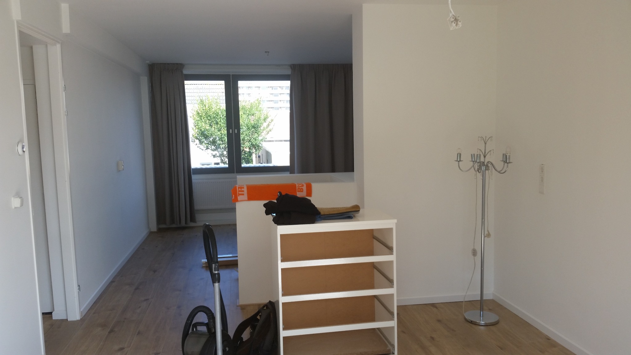 Pusty, jasny pokój po remoncie z parkietem, zasłonami w oknach, lampą podłogową i regałem bez półek, przygotowany do umeblowania.