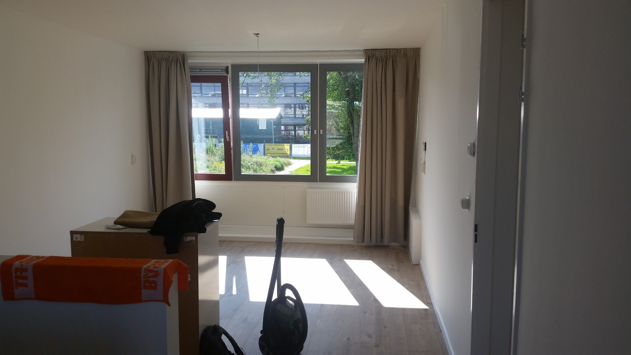 Pusty, jasny pokój po remoncie z drewnianą podłogą, zasłoniętymi oknami i odkurzaczem na podłodze, przygotowany do umeblowania.